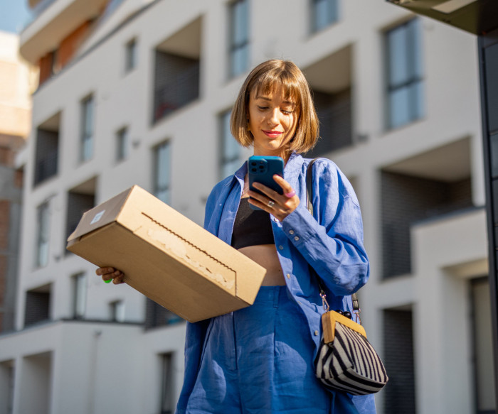 Une femme tient un colis dans une main et un smartphone dans l'autre main.